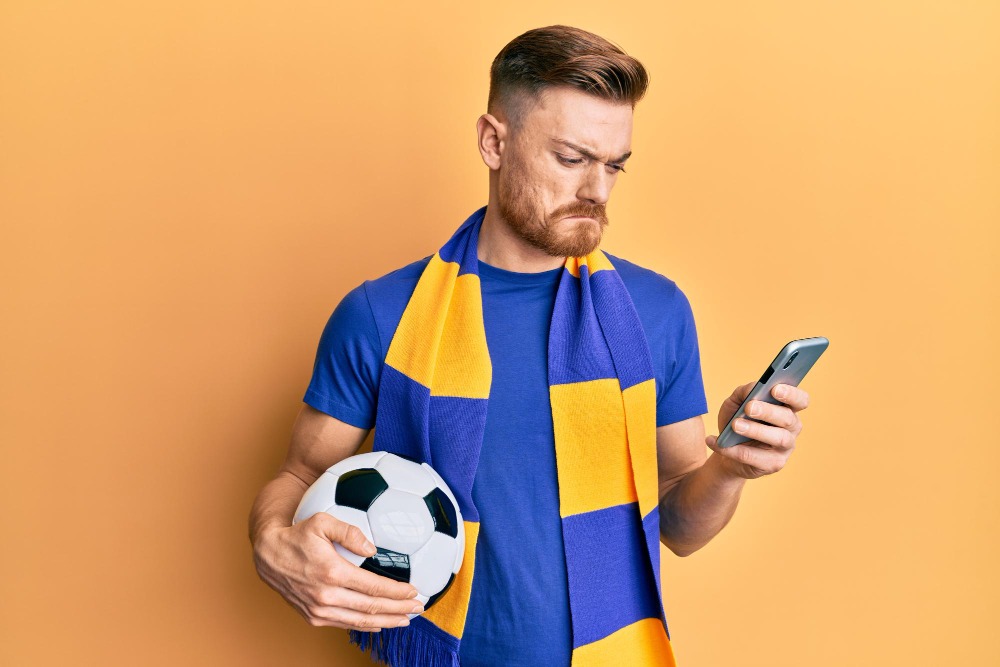 Homem com bola preocupado olhando para celular - Reprodução: Imagem de krakenimages.com no Freepik